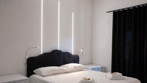 A neatly made bed with white linens and two pillows, a bedside table with a lamp, and a window with black curtains. - B&B Black & White (Pietramontecorvino)