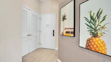 Um corredor com uma porta branca, um quadro com uma ilustração de abacaxi e um quadro com uma cena de praia.