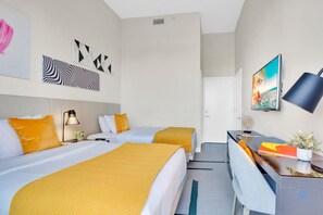 A hotel room with two beds, a desk, a chair, a desk lamp, and artwork on the walls.