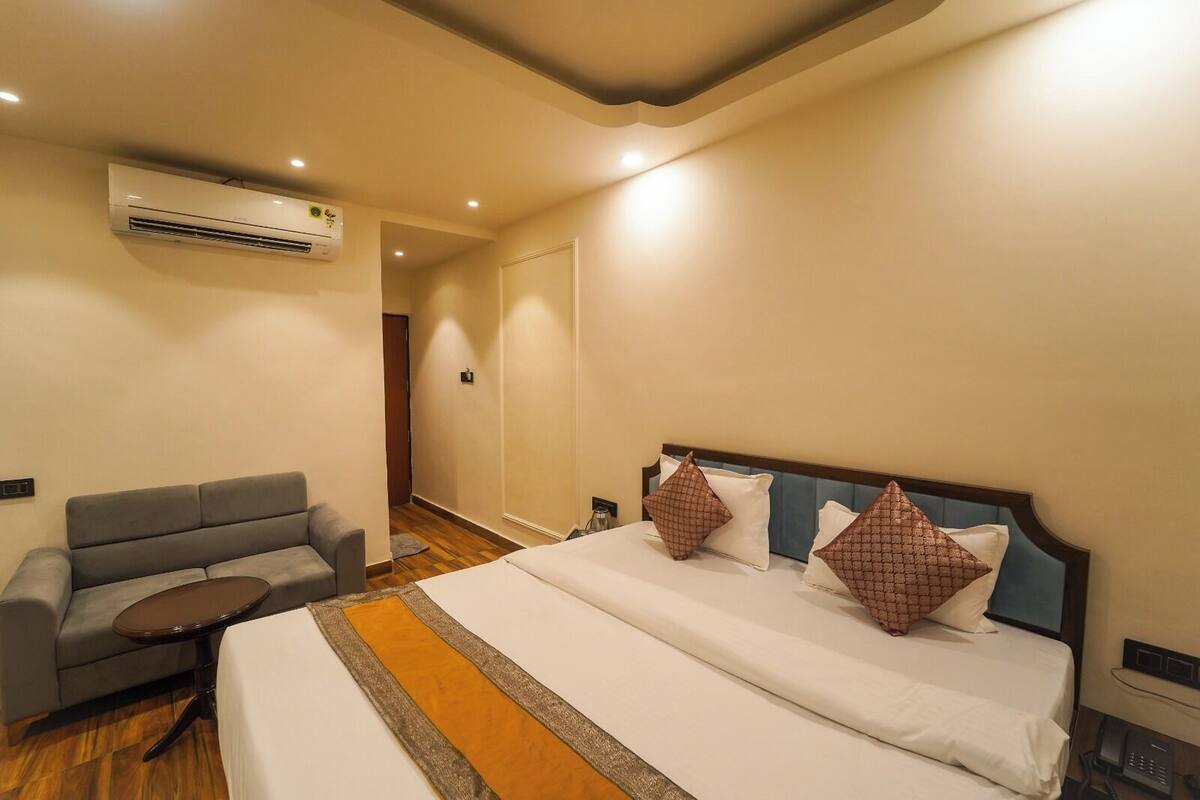 A hotel room with a bed, a sofa, a small table, and an air conditioning unit.