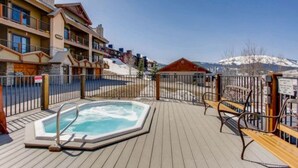 Een houten terras met een hottub, lounge stoelen en uitzicht op besneeuwde bergen.