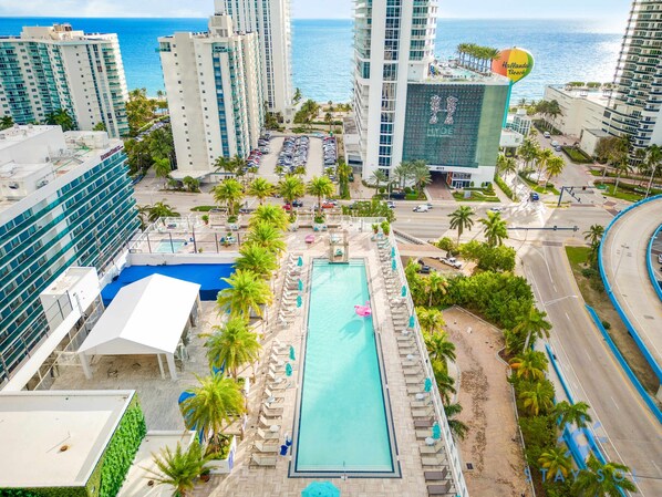 A hotel with a swimming pool surrounded by palm trees and lounge chairs, overlooking a beach and ocean.