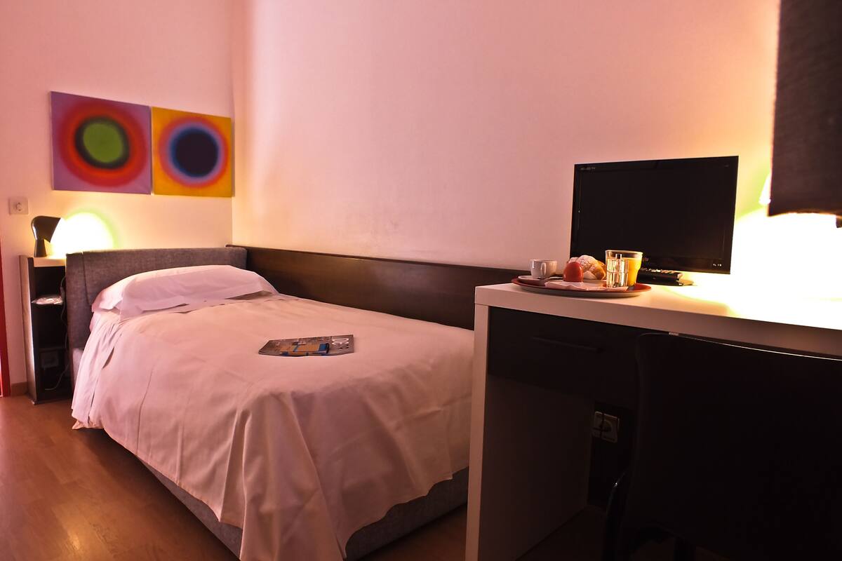Une chambre d’hôtel avec un lit simple, un bureau avec un ordinateur, une lampe et deux tableaux abstraits accrochés au mur.