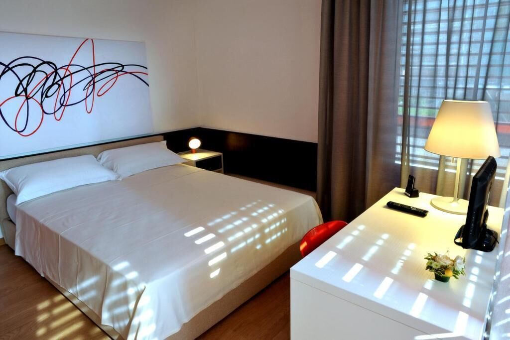 A hotel room with a bed, bedside table, lamp, and abstract art on the wall.