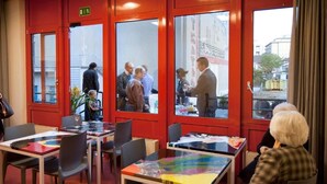 Een restaurant met rode muren en grote ramen, tafels met kleurrijke tafelkleden, en mensen die er dineren.