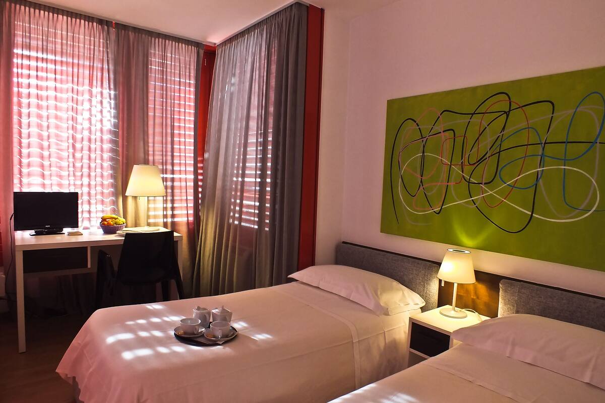 Une chambre d’hôtel avec deux lits, un bureau avec un ordinateur et un grand tableau abstrait accroché au mur.