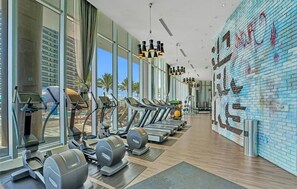A gym with treadmills, elliptical machines, and weight stations, large windows, and a brick wall mural.