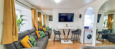 A modern living room with a grey sofa set, a dining table with two chairs, a TV mounted on the wall, and a washing machine in the corner.