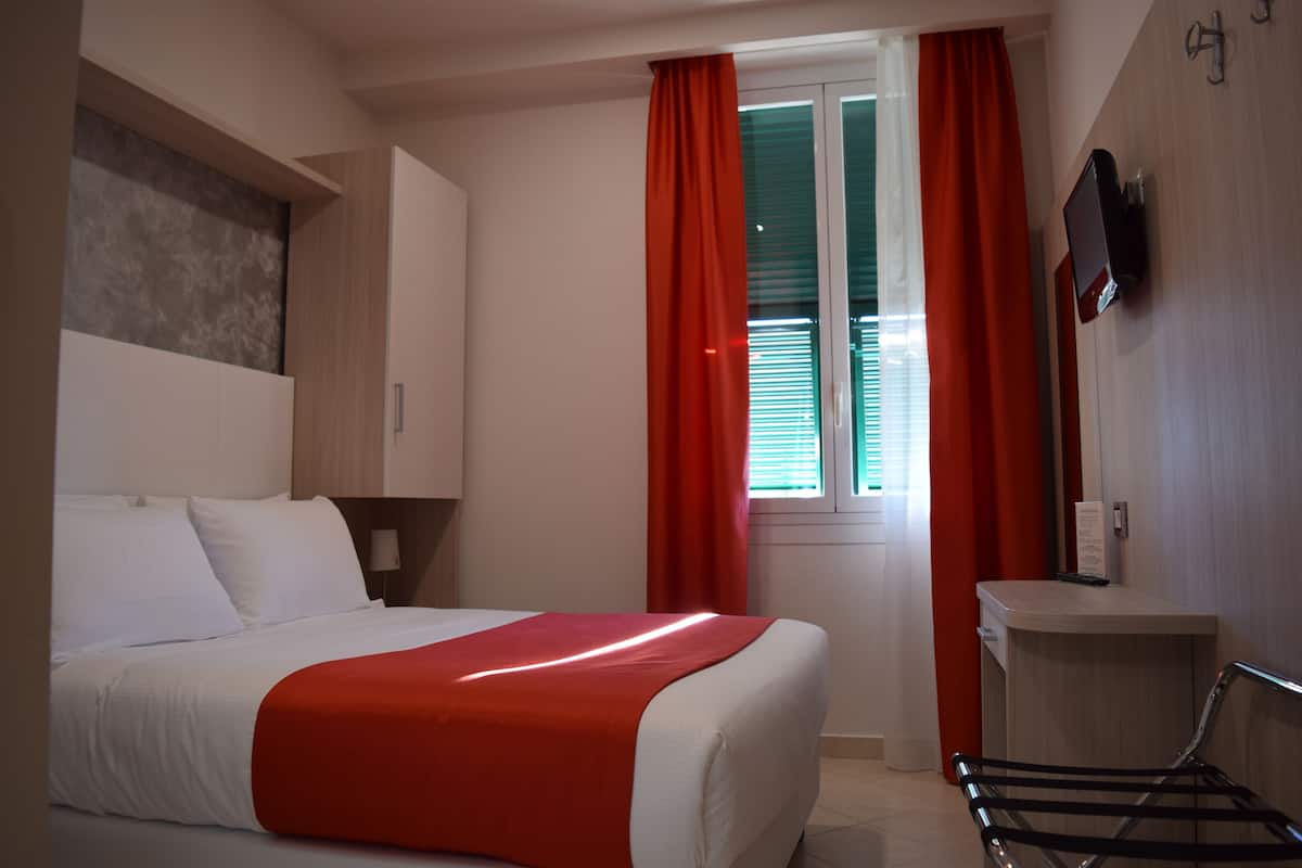 Una habitación de hotel con cama, escritorio, televisión y ventana con cortinas.