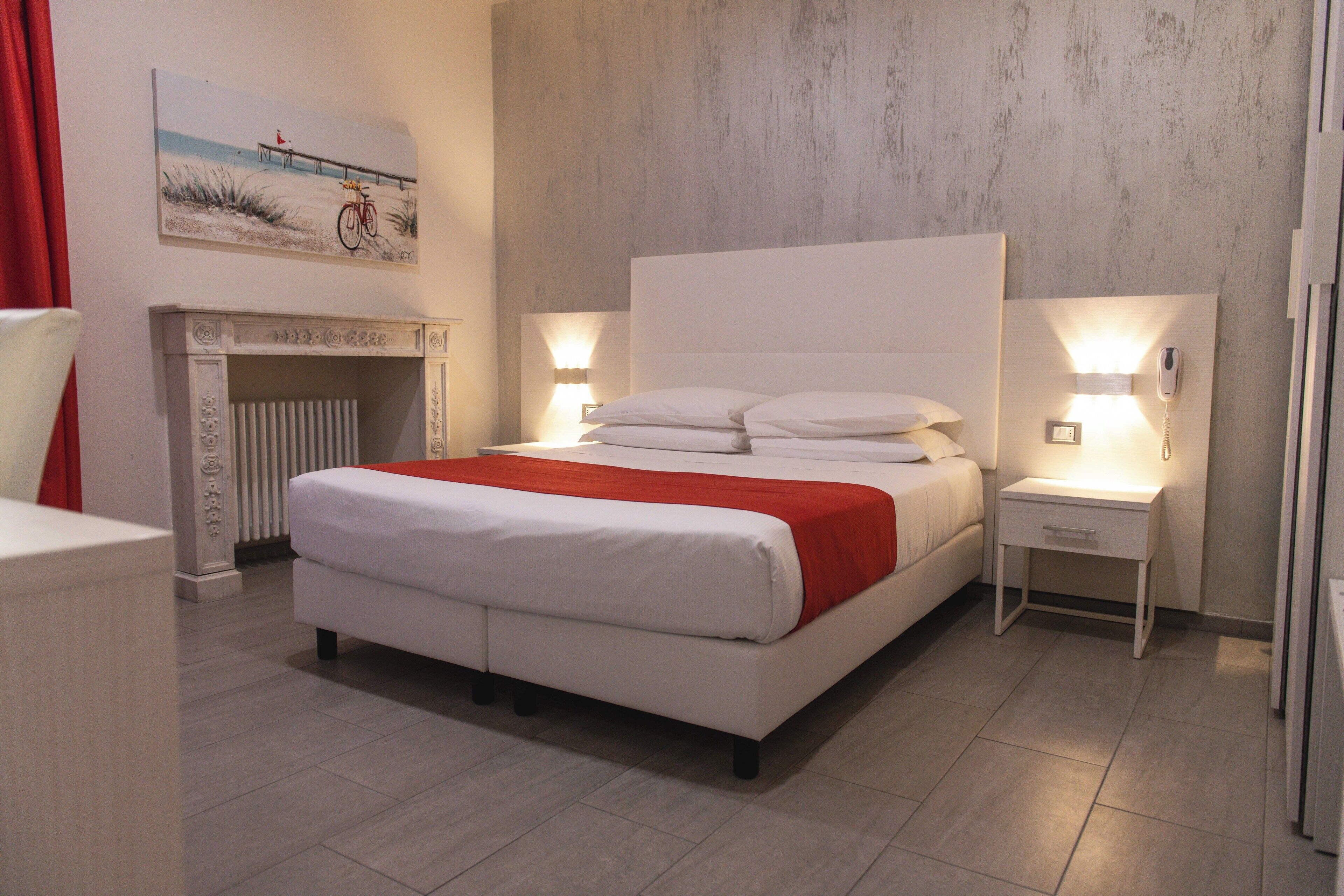 A modern bedroom with a large bed, bedside tables with lamps, a fireplace, and a beach-themed painting on the wall.