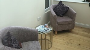 A modern living room with a glass ceiling, a grey armchair, a smaller armchair with a cushion, a small round table with newspapers, and a painting on the wall.
