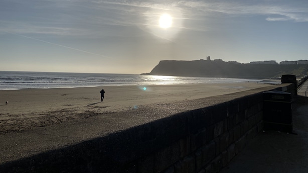 A beach with a stone wall, ocean waves, and a distant cliff with buildings.