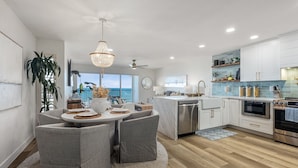A modern kitchen with white cabinets, a dining area with grey chairs, and a large window overlooking the ocean. - Del Mar Beach Club Condo with Ocean Views & Pool (Solana Beach)