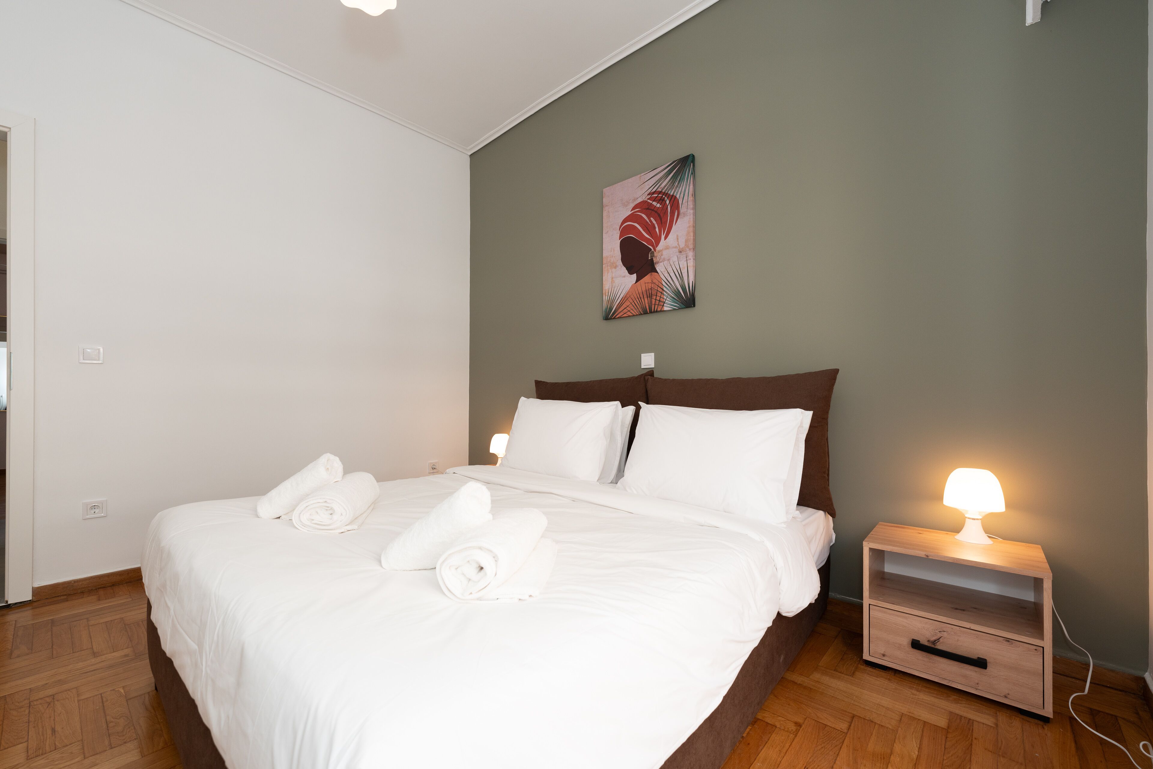 A neatly made bed with white linens and rolled towels, a bedside table with a lamp, and a painting on the wall.
