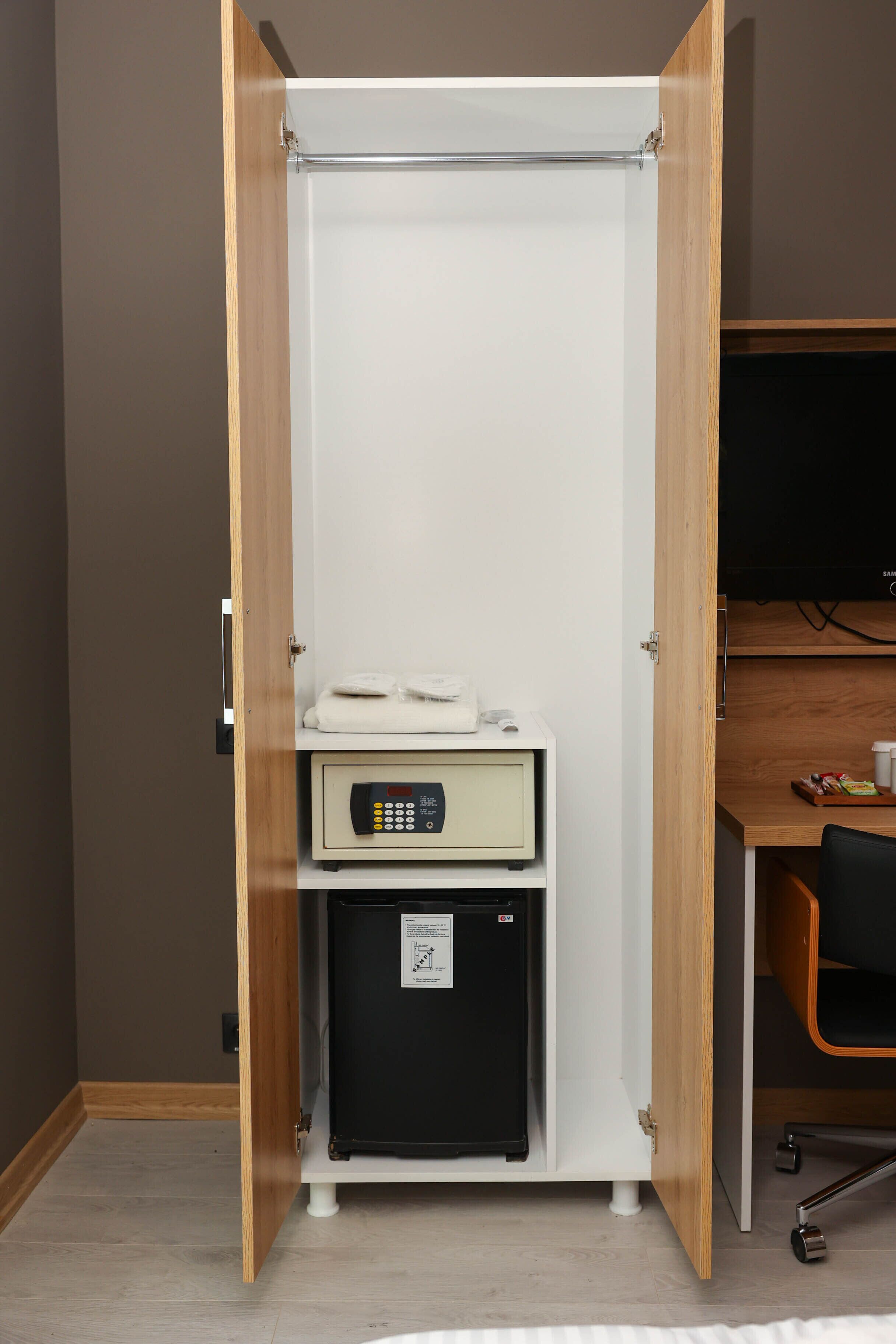A room with a wooden wardrobe, a small black refrigerator, a control panel, and a desk with a chair.