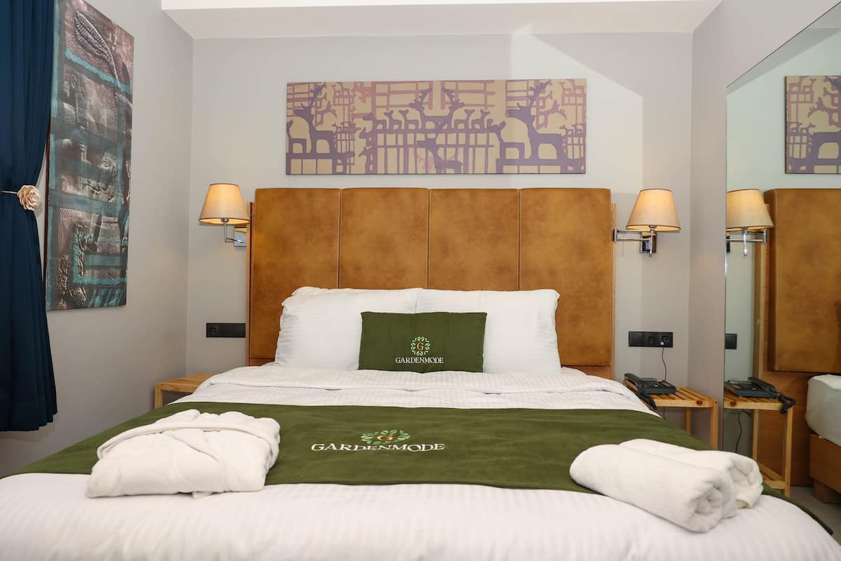 Una habitación de hotel con una cama grande, dos lámparas de noche, un cabecero, una toalla verde y una almohada con el logotipo "GARDENMODE".