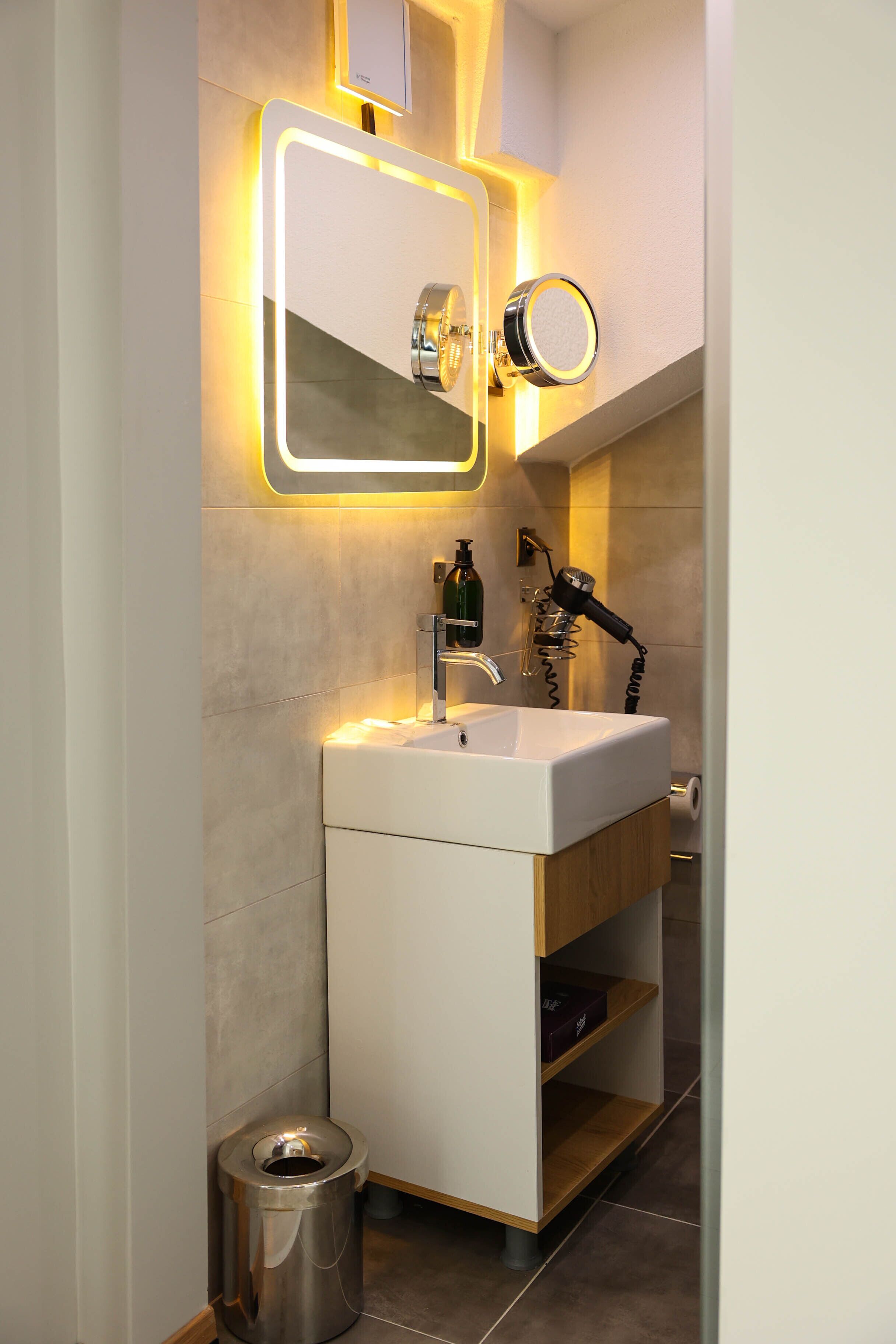 Un baño moderno con un lavamanos blanco, un espejo con iluminación perimetral, un secador de pelo y un cubo de basura.