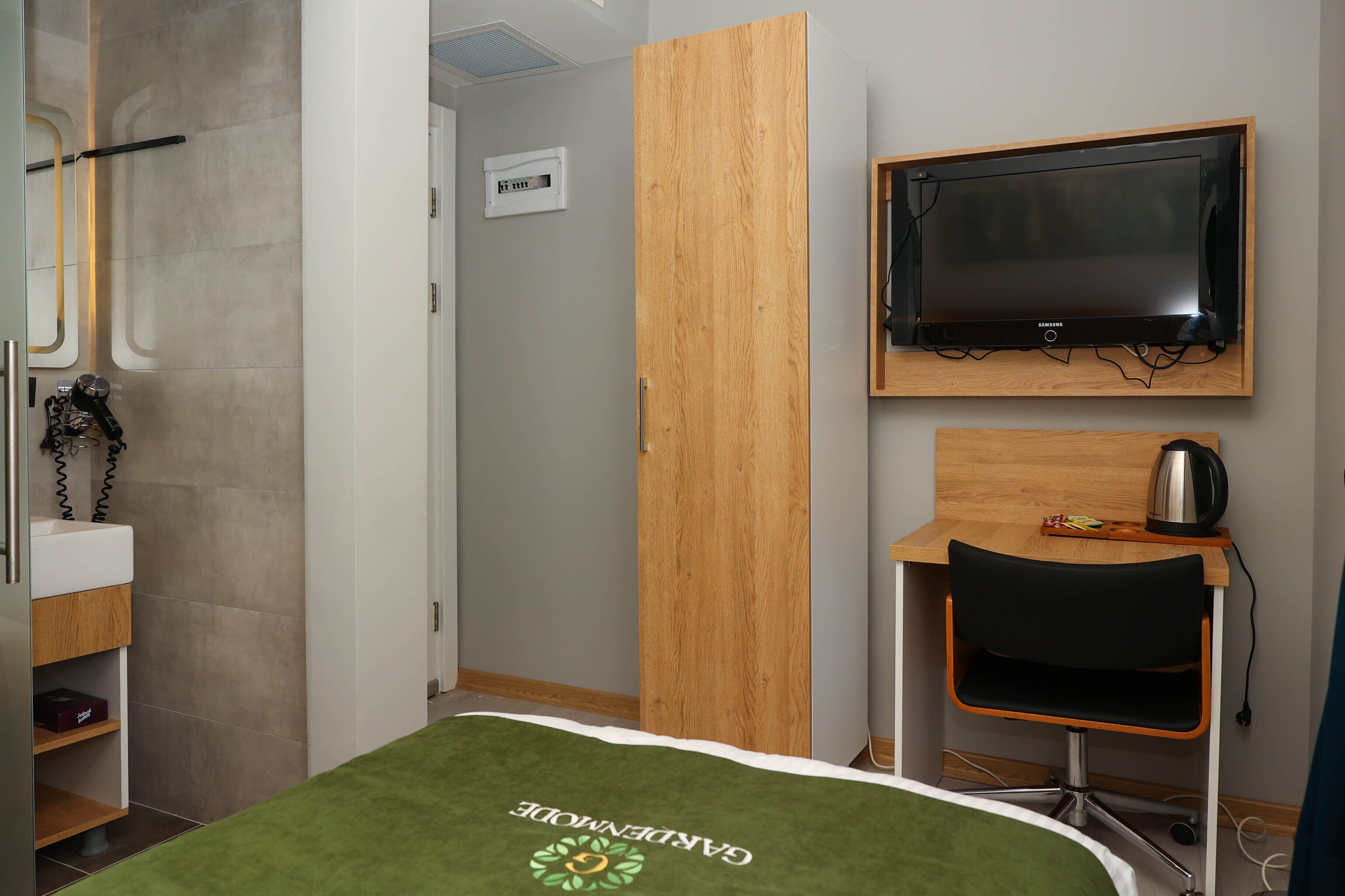A hotel room with a bed, a TV mounted on the wall, a desk with a chair, and a wooden wardrobe.