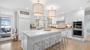 A modern kitchen with white cabinetry, a large island with bar stools, pendant lighting, and a view of the outdoors. - The Ultimate Cape Cod luxury home. Dock, pool, hot tub and views! RPMS104 (Osterville)