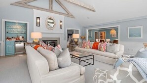 A spacious living room with two sofas, a coffee table, and a fireplace. There is a kitchen area with cabinets and a dishwasher. The room has a high ceiling with exposed beams and a large mirror above the fireplace. - The Ultimate Cape Cod luxury home. Dock, pool, hot tub and views! RPMS104 (Osterville)
