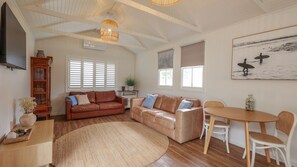 A spacious living room with a leather sofa, a dining table with chairs, a large window with blinds, and a painting on the wall.