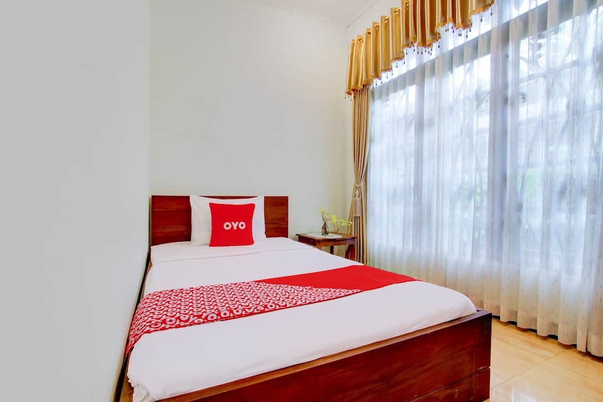 Ein Hotelzimmer mit einem hölzernen Bett, einer roten und weißen Bettdecke, einem roten Kopfkissen mit dem Aufdruck "OYO" und transparenten Vorhängen mit einem goldenen Wadenstreifen.
