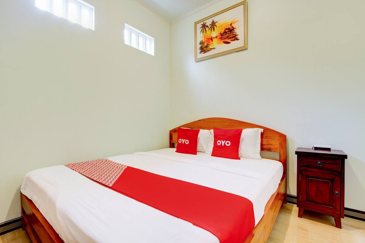 Ein ordentlich gemachtes Bett mit roter Decke und Kissen, ein Holz-Kopfstück, ein Nachttisch mit Schublade und ein Bild an der Wand.