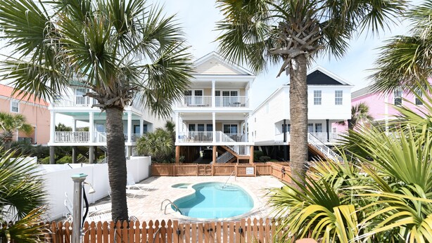 A pool area with a wooden fence, palm trees, and a two-story building with balconies. - Oceanfront 5 BR Beach House with a Private Pool! 19th Hole (Surfside Beach)