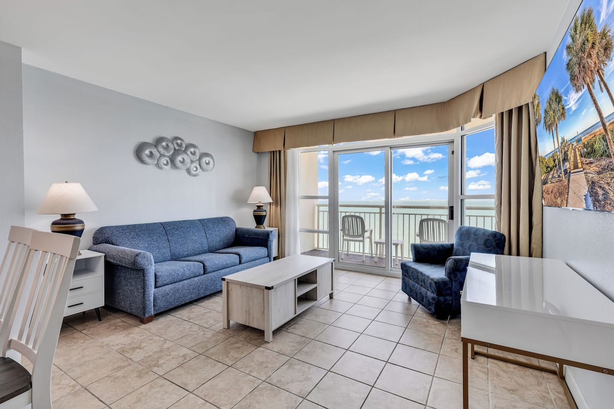 Un salon moderne avec un canapé bleu, une table basse blanche et une table à manger. Il y a un balcon avec vue sur les palmiers et l’océan.