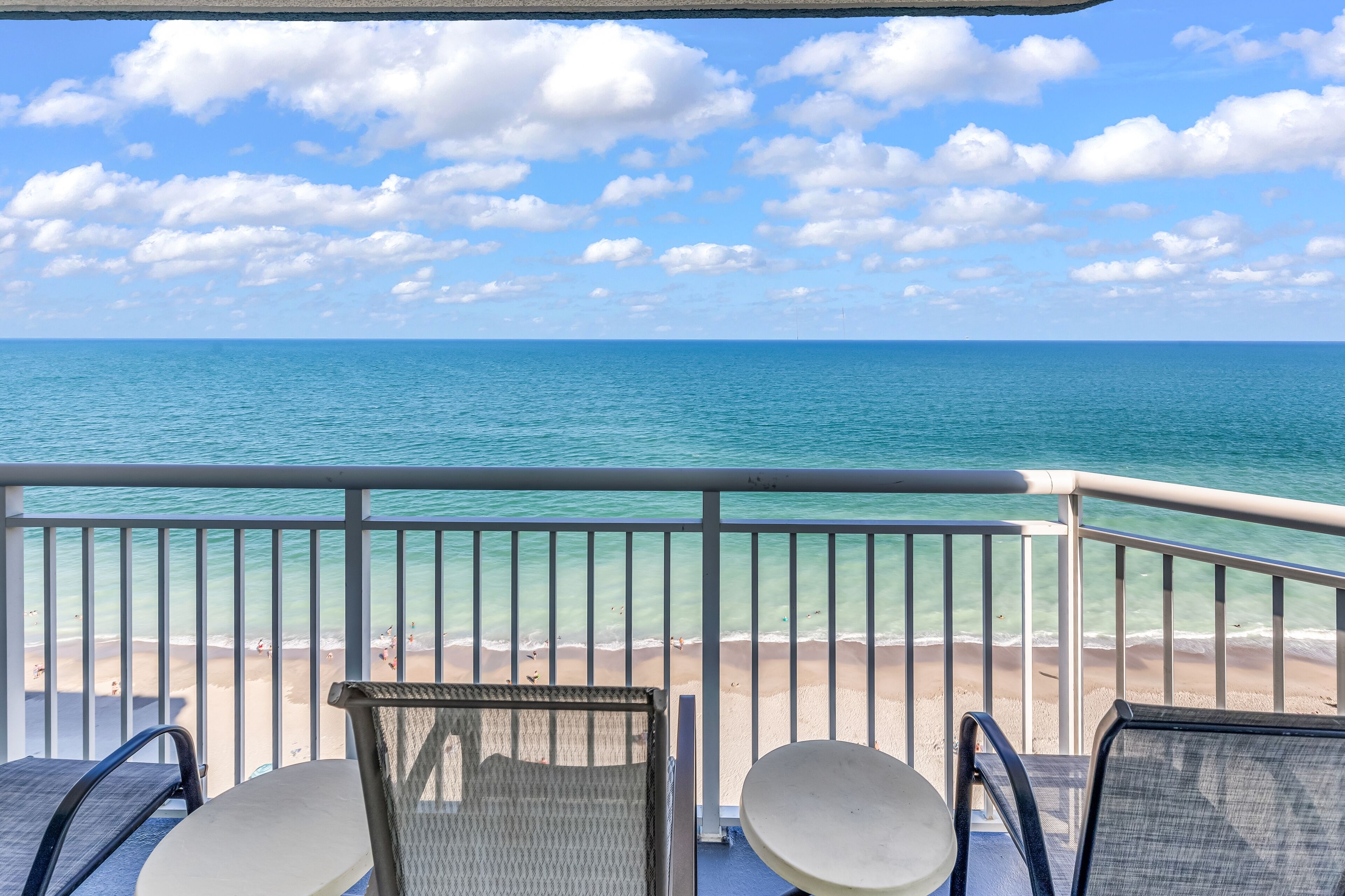Un balcon avec deux chaises et une table, donnant sur une plage aux eaux turquoise et limpides et au rivage sablonneux.