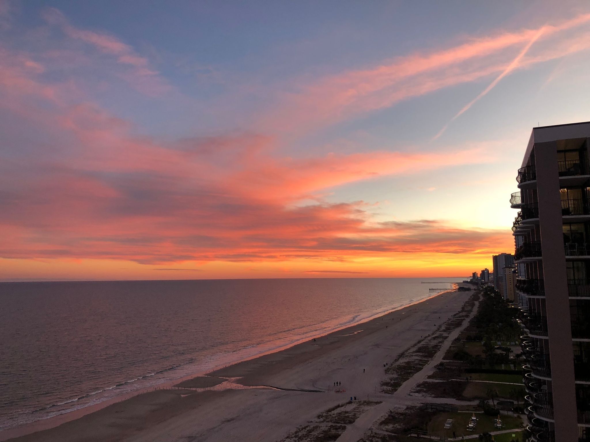 Blick auf einen Strand bei Sonnenuntergang, mit Gebäuden und einem Steg.