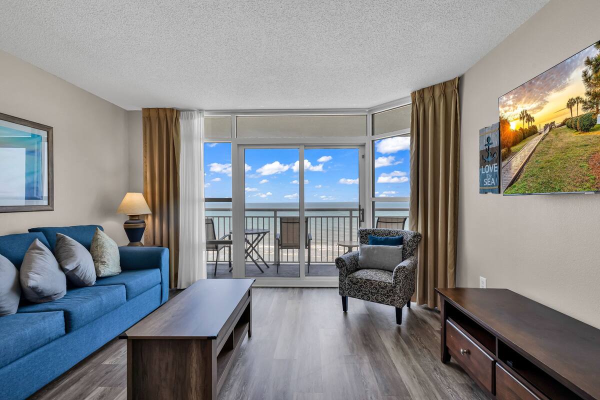 Une chambre d’hôtel moderne avec un balcon, un canapé, un fauteuil, une table et un tableau.