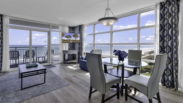 A modern living room with a glass table, chairs, a TV, and a view of the ocean. - Carolinian Resort (Myrtle Beach)