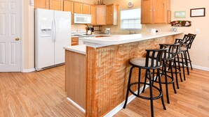 A kitchen with wooden cabinets, a white refrigerator, a microwave, and a sink. There are four black bar stools around the island. - Walk to the Beach 5 BR Beach House with a Private Pool! Catching R.A.E.S (Surfside Beach)