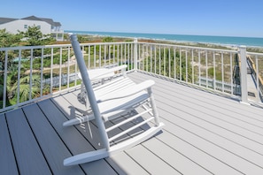 A wooden deck with a rocking chair, white railings, and a view of the beach and ocean.