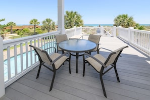 A covered outdoor area with a round table and chairs, a white railing, and a view of the ocean.