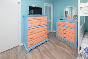 A bedroom with two peach-colored dressers, a television on the left dresser, a mirror on the right, and a bed with a patterned bedspread.