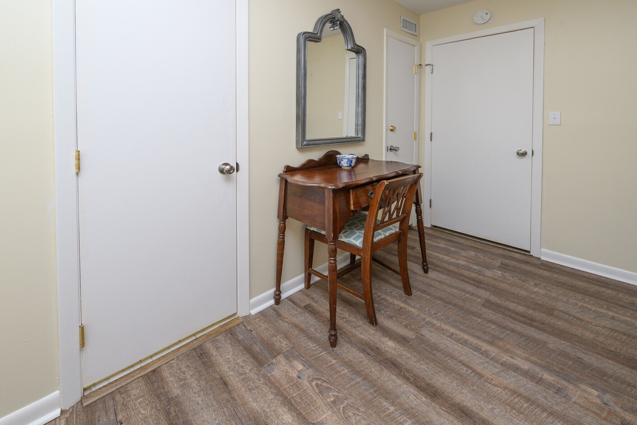 A room with a wooden desk, a chair, a mirror, and two white doors.
