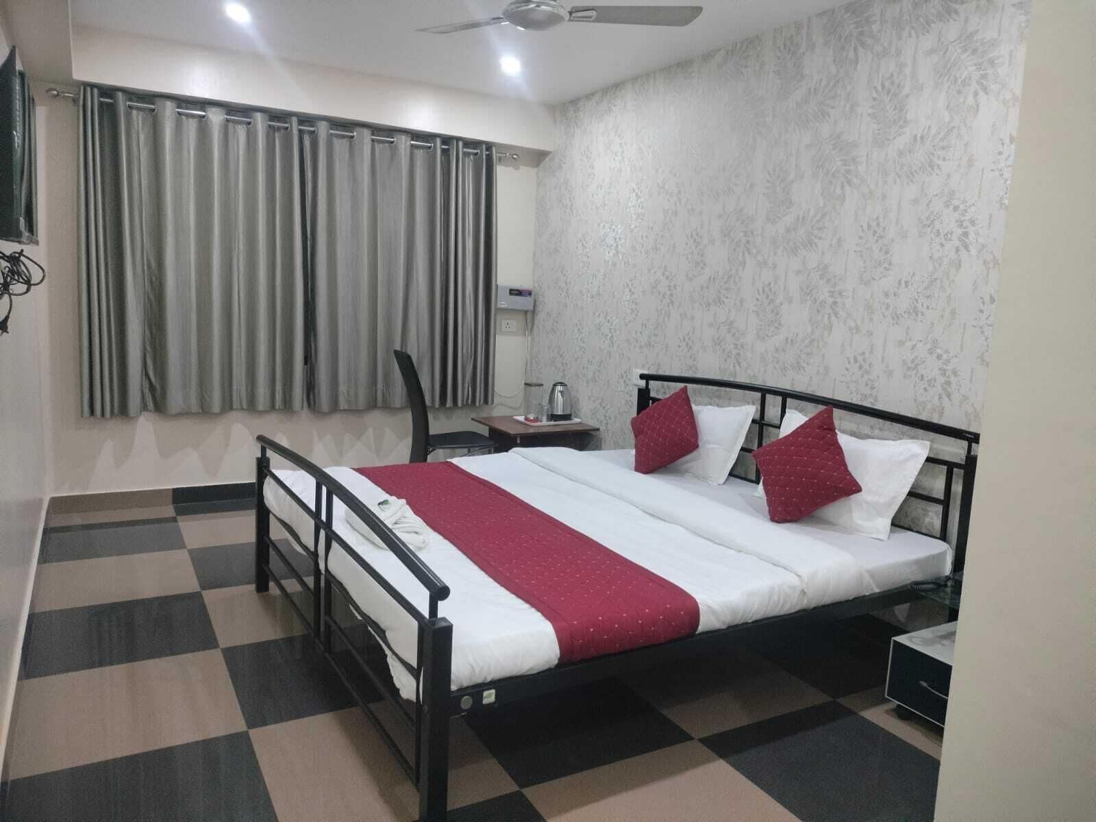 A hotel room with a checkered floor, a bed with white and red bedding, a desk with a kettle, and a window with curtains.