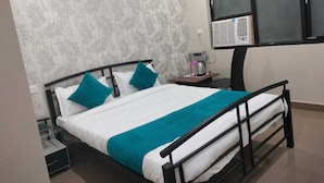 Een netjes opgemaakt bed met een blauwgroene sprei en kussens, een klein nachtkastje met een waterkoker, en een raam met een airconditioning.'