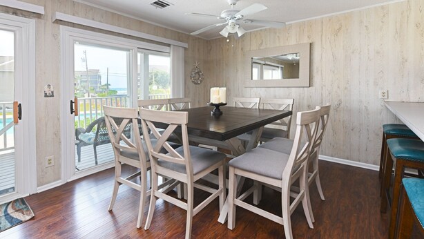Un coin repas avec une table en bois, six chaises, un ventilateur de plafond et une vue sur l’extérieur grâce à de grandes portes-fenêtres coulissantes.