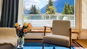 A room with a view of mountains, a chair, a table, and a vase with flowers.