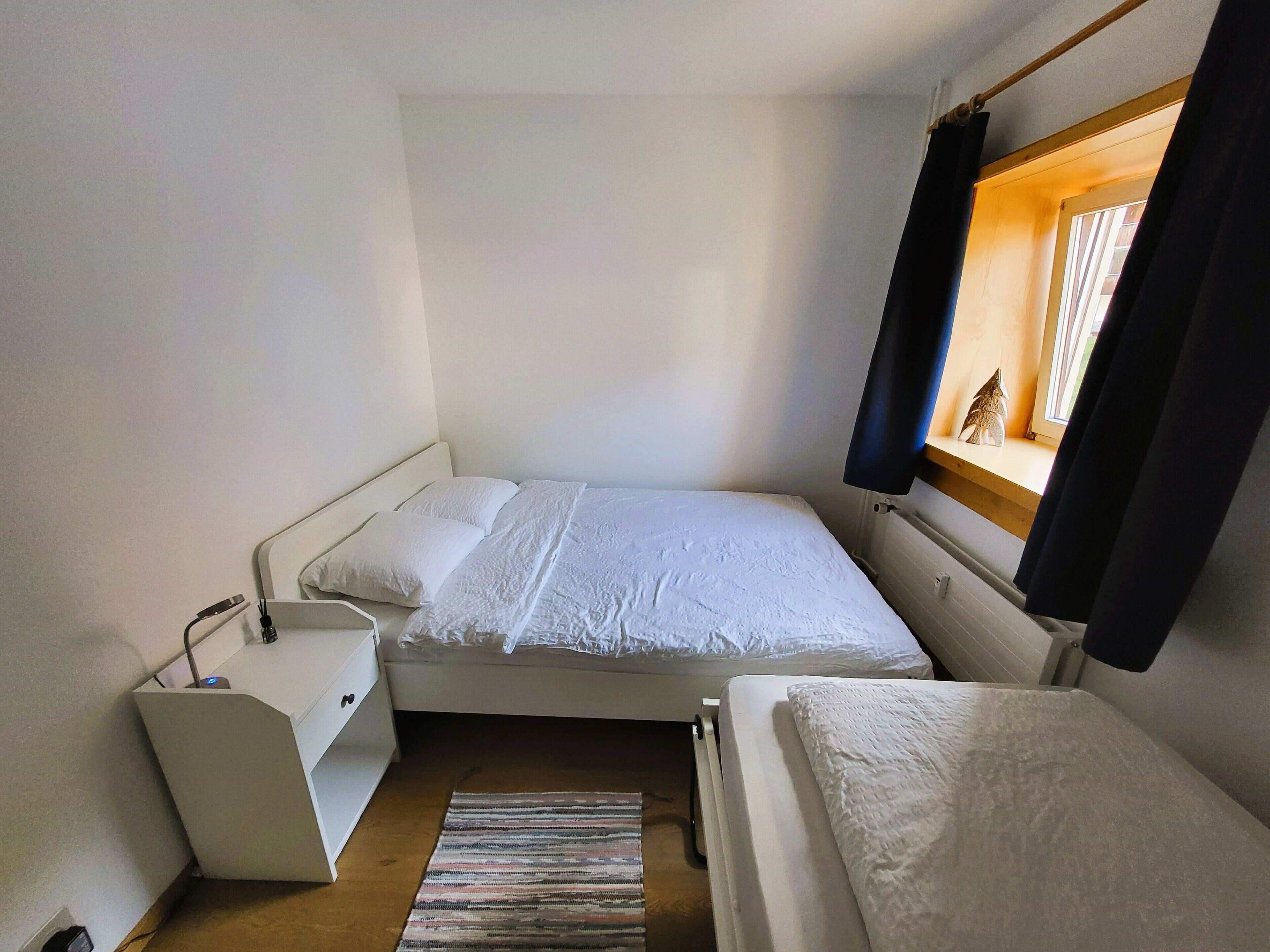 A small, single-bed room with a white bed, a bedside table, and a window with curtains.