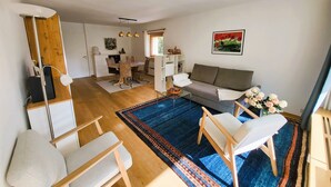 A modern living room with a sofa, chairs, a coffee table, and a dining area with a table and chairs. There is a large rug on the floor, and a painting hangs on the wall.