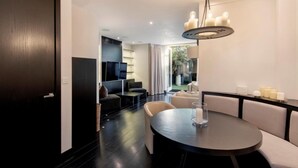 Une chambre d’hôtel moderne avec une porte en bois foncé, une table à manger ronde entourée de chaises, une grande fenêtre avec des rideaux et un lustre.