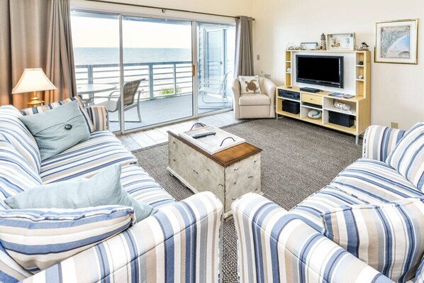 A living room with striped sofas, a coffee table, a TV stand, and a balcony with a view of the sea.