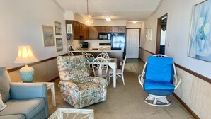 A compact living space with a sofa, a patterned armchair, a blue chair, a dining table with chairs, a kitchen area, and a hallway leading to a door. - Oceanfront 2 BR Condo with a Shared Pool! Sea Shadow 402   (Surfside Beach)