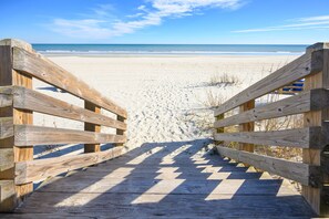 Une promenade en bois mène à une plage de sable fin, sous un ciel bleu et dégagé.