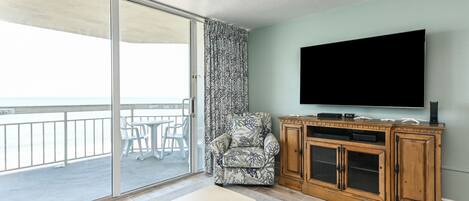 A living room with a large TV, a wooden cabinet, a patterned armchair, and a sliding glass door to a balcony.