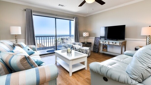 Un salon spacieux doté d’une grande fenêtre donnant sur la plage, d’un téléviseur à écran plat et d’un coin salon confortable avec des canapés à rayures et une table basse.